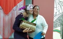 Two women hug while one holds a basket with a flower.