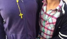 A yellow-gold rosary sits on a woman's purple jacket. Next to her is another person with a green rosary around their neck.